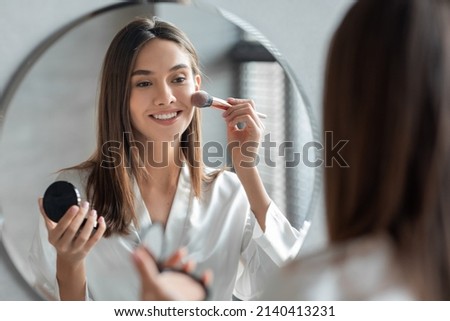 Attractive Young Woman Doing Daily Makeup While Standing Near Mirror In Bathroom, Happy Beautiful Female Putting Blush On Cheeks While Getting Ready At Home, Selective Focus On Reflection Stockfoto © 