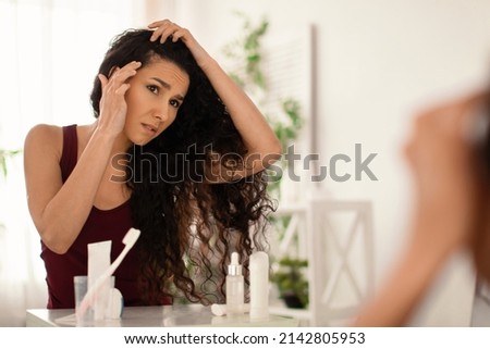 Frustrated millennial female having dandruff or grey hair problem, taking care of herself at home, copy space. Anxious young woman looking at her roots with flaky scalp in mirror indoors Foto d'archivio © 