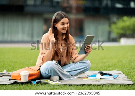 Cheerful pretty brunette young woman in casual university student chilling at park, sit on grass, doing homework, using digital tablet, have video call with friends, empty space. Telecommunication Imagine de stoc © 