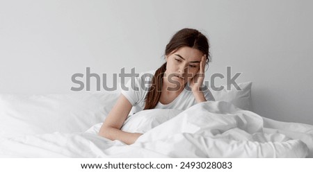 Sad european millennial female, worried, suffers from insomnia and headache. Lady sits on bed in bedroom at home, copy space. Migraines, health and mental problems, lack of sleep and depression