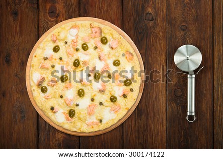 Delicious seafood pizza with salmon, capers and olives - thin pastry crust isolated at wooden background with stainless steel cutter, above view