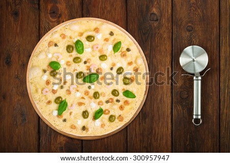 Delicious seafood pizza with shrimps, calamari rings, capers and olives - thin pastry crust at wooden table background with stainless steel cutter, above view
