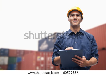 Smiling young engineer in protective work wear in shipping yard examining cargo