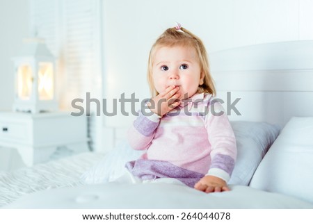 Funny surprised blond little girl sitting on bed in bedroom. White interior, big bed, bedside table and night lamp. Girl touches her mouth by hand