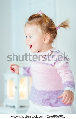 Excited blond little girl with ponytail jumping on bed, laughing and screaming. White interior, bedroom, night lamp