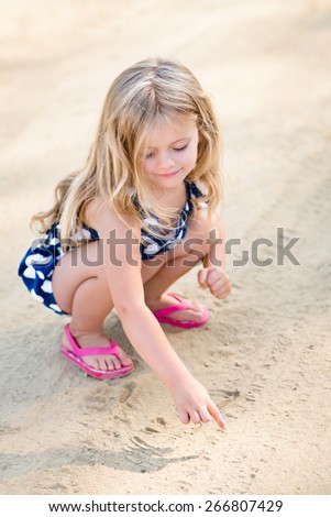 Sweet smiling little girl with long blond hair squatting and drawing in the sand on the beach in summer day