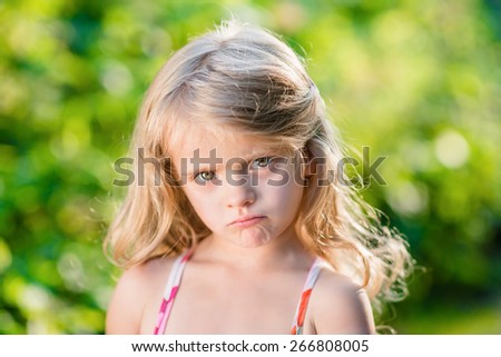 Close-up portrait of sad blond little girl with pursed lips. Sunny summer day in beautiful park