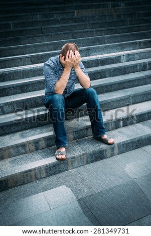 Outdoor portrait of despaired young man covering his face with hands sitting on stairs. Feelings of sadness, despair and tragedy