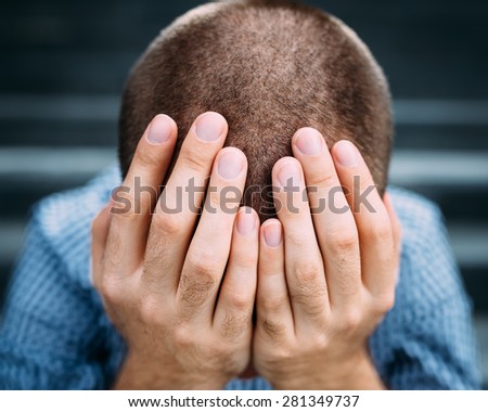 Closeup portrait of despaired young man covering his face with hands. Selective focus on hands. Sadness, despair, tragedy concept