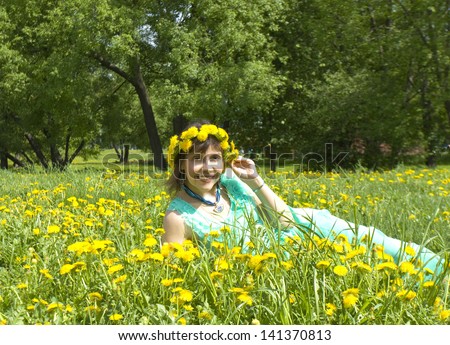 Pretty lady in wreath of yellow dandelions brunette European, laying on green meadow with many yellow dandelions in park.