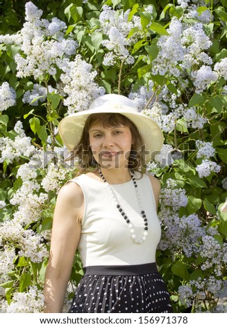 European pretty woman brunette in white hat half body near shrub of white lilac.