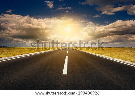 Driving on an empty new asphalt road through the agricultural yellow fields towards the setting sun.