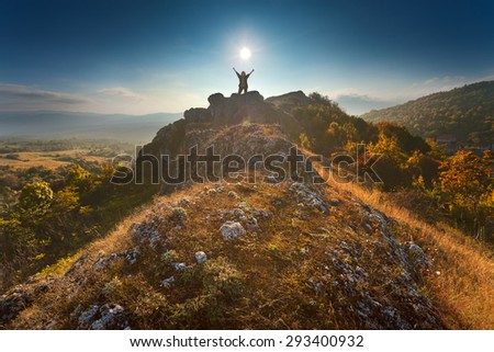 Woman hiker standing on a rock cheering elated with arms raised in the sky towards the setting sun with colorful valley in background. Success victory concept.