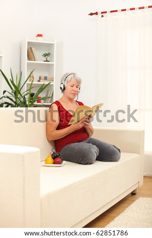 Senior woman sitting on sofa, reading book and listening to music