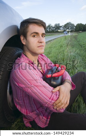 young man sitting on the front wheel of a car with a black petrol cans and perplexed looks into the camera
