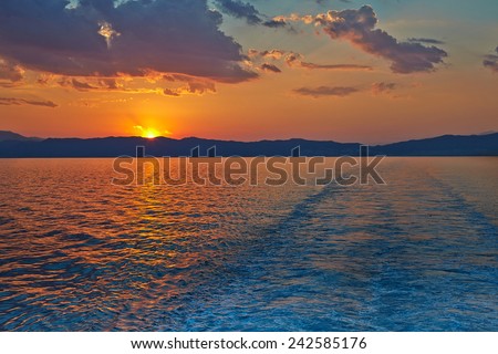 Sunset over the Greece, waves from the ferry, and the Mediterranean sea. The sun sets behind the mountains on the horizon.