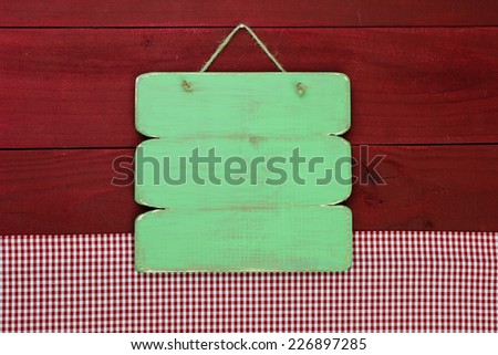Blank antique green rustic sign by red checkered tablecloth hanging on red wooden board background