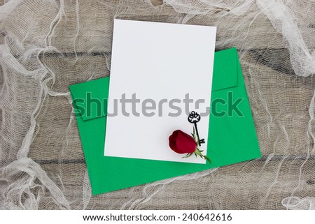 Blank white letter or note card and green envelope with red flower and black iron key on shabby netting background