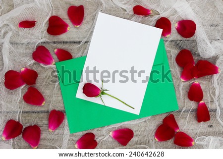 Blank white letter or note card and green envelope with red flower and red rose petals on shabby white netting and wood background