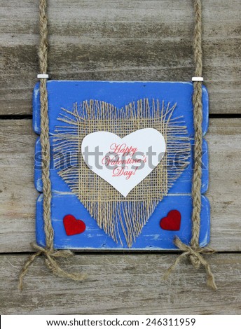 Happy Valentine\'s Day text on white and burlap hearts and red hearts on antique blue wood sign hanging by braided rope on rustic wooden background