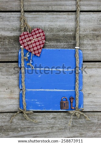 Blank antique blue sign with red checkered heart, lock, and key hanging by braided rope on rustic wood background