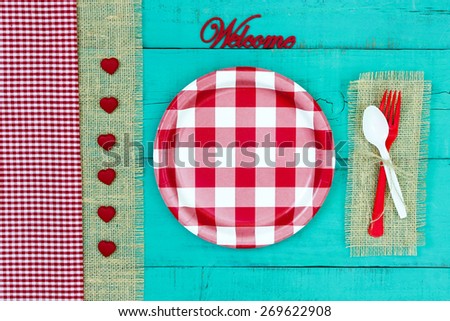 Welcome sign with red and white checkered plate, hearts and gingham and burlap border on antique teal blue wood background; above view looking down