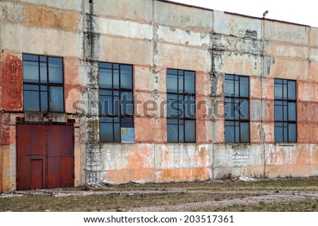 Big abandoned old factory building with doors and windows closed . Dirty wall with cracked plaster leafless