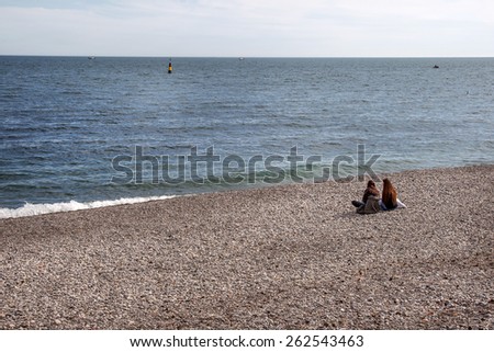 Two girls looking into the distance and communicate sitting on the beach on the pebble stone desert autumn beach waiting for the summer heat and sun. Overcast autumn day