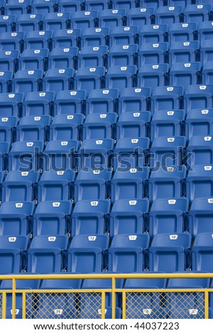 Blue seats with a yellow metal fence in front of them, at a stadium. These seats are numbered and make an ideal background.