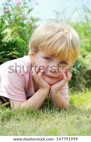a sweet young child lays in the grass outside by flowers, smiling and resting his head in his hands