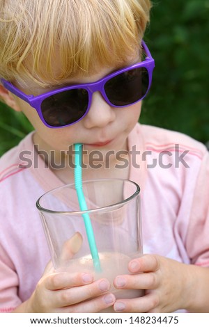 A young child wearing sunglasses is holding a big glass of chocolate milk and sipping it through a straw on a sunny summer day