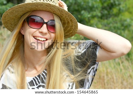 a young, attractive woman with long blonde hair, is sitting outside on a sunny summer day, wearing a straw hat and sunglasses