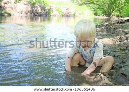 a young boy child is sitting in on the beach of a river in the woods, playing in the mud on a summer day.