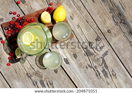 A pitcher full of fresh squeezed lemonade is sitting on a wood picnic table background, surrounded by red raspberry fruit on a summer day.