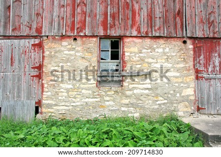 A broken window is seen between two wooden doors with peeling red painted barnwood, on an old abandoned barn.
