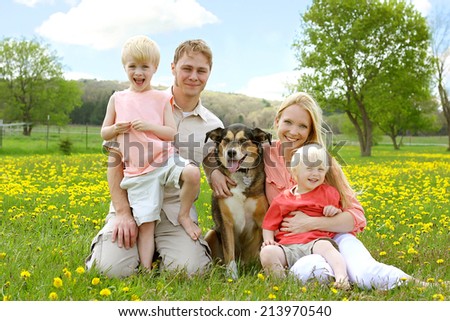 A happy family of four people, mother, father, young child and toddler are sitting in a meadow of Dandelion flowers with their German Shepherd mix dog, on a beautiful Spring day.