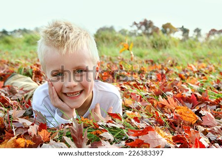 A portrait of a happy, smiling five year old child, laying outside in the fallen yellow and red Maple Tree leaves on an Autumn day.