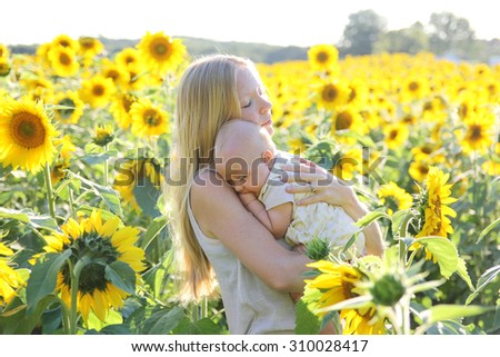 A happy young mother is lovingly holding her baby daughter outside in a sunflower flower field in the summer.
