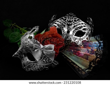 Masks with rose.Silver carnival masks laying with folding fan and red rose on black fabric background.