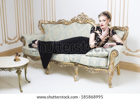 Beautiful blonde royal woman laying on a retro sofa in gorgeous luxury dress. Indoor. Copy Space