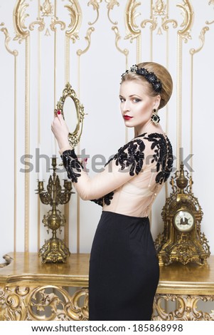 Beautiful blonde royal woman standing near retro table in gorgeous luxury dress with mirror in her hands. Indoor