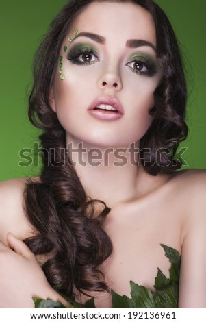 Beautiful brunette dryad woman with creative make up and beads on her face, curly hair and costume made of leaves on green background