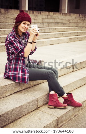 Brunette woman in hipster outfit sitting on steps and photographing on retro camera on the street. Toned image