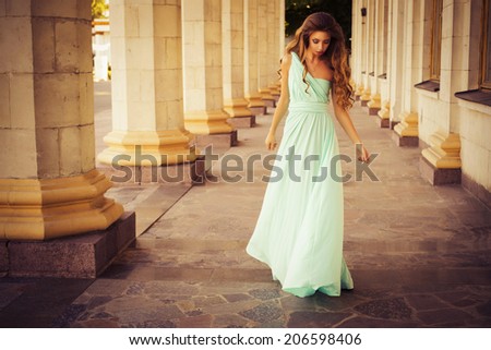 Beautiful blonde with a long curly hair in a long evening dress in motion outdoors near retro vintage building in summer sunset. Copy Space