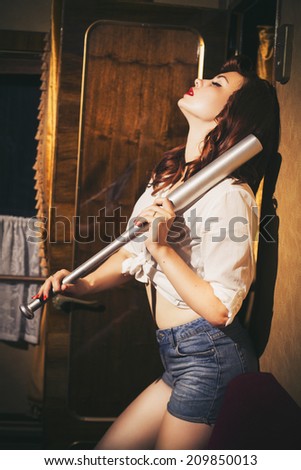 Beautiful sexy brunette retro woman with pin-up hairstyle and make-up in a shorts and shirt with a bat in her hands in vintage interior. Indoors. Retro toned