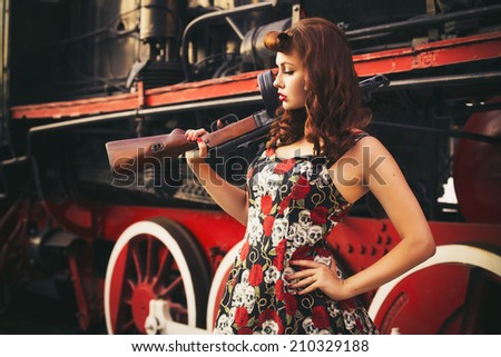 Beautiful sexy brunette retro woman with pin-up hairstyle and make-up in a shorts and shirt with a gun in her hands on vintage train. Outdoors. Retro toned