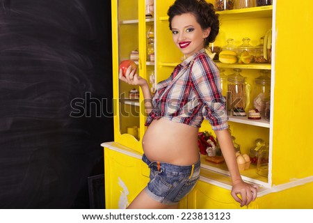Beautiful brunette woman on a 7th month pregnancy in plaid shirt and jeans shorts on a yellow kitchen and charcoal board. pin up style. copy space