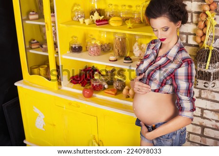 Beautiful brunette woman on a 7th month pregnancy in plaid shirt and jeans shorts on a yellow kitchen. pin up style. copy space
