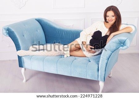 Beautiful brunette woman on a 7th month pregnancy in white sweater. light interior. blue sofa. copy space