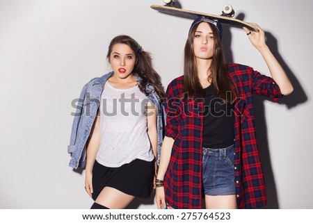 Two beautiful brunette women (girls) teenagers spend time together having fun, make funny faces. Casual hipster outfit, jeans jacket and plaid shirt, with a skateboard